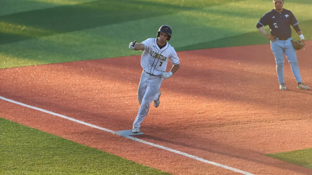 Baseball runner in a white uniform slides into a base on a sunlit red infield as a fielder watches in the background.