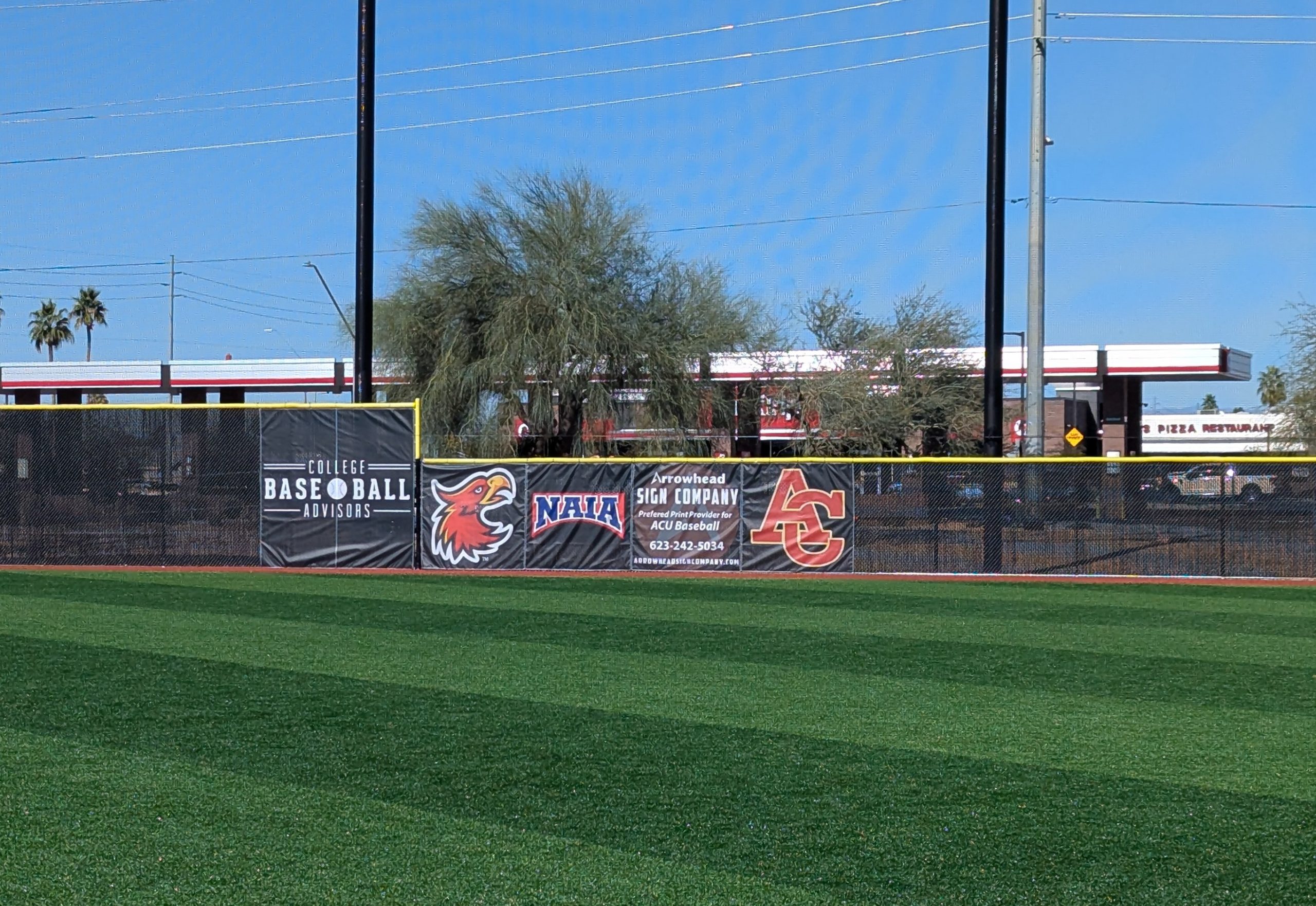 Baseball field outfield fence with advertising banners and logos, blue sky in background.