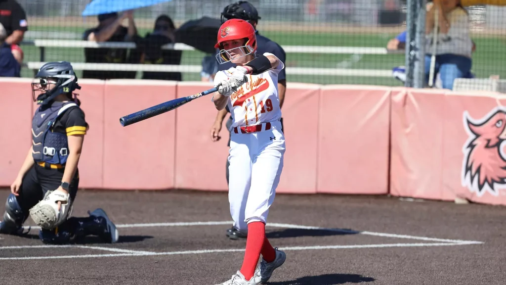 Baseball batter in red helmet and white uniform swings at a pitch as the catcher watches from behind in protective gear.