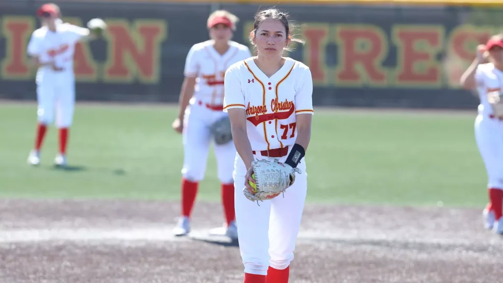 Female softball player in white and red uniform stands on the infield, glove cupping a ball, facing the camera.