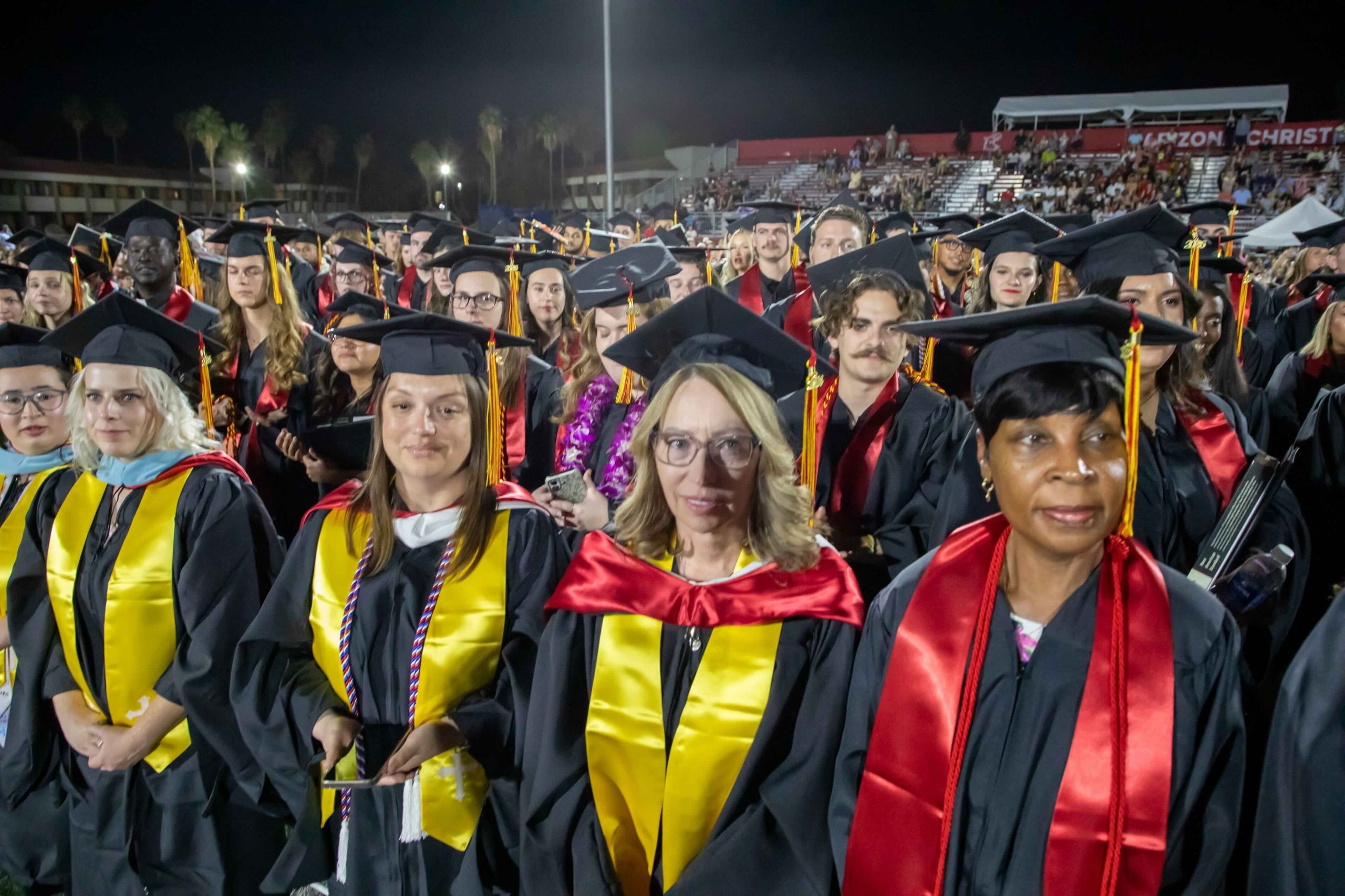 Crowd of graduates in caps and gowns at a nighttime stadium ceremony, many wearing yellow stoles and red sashes, past rows of bleachers in the background.
