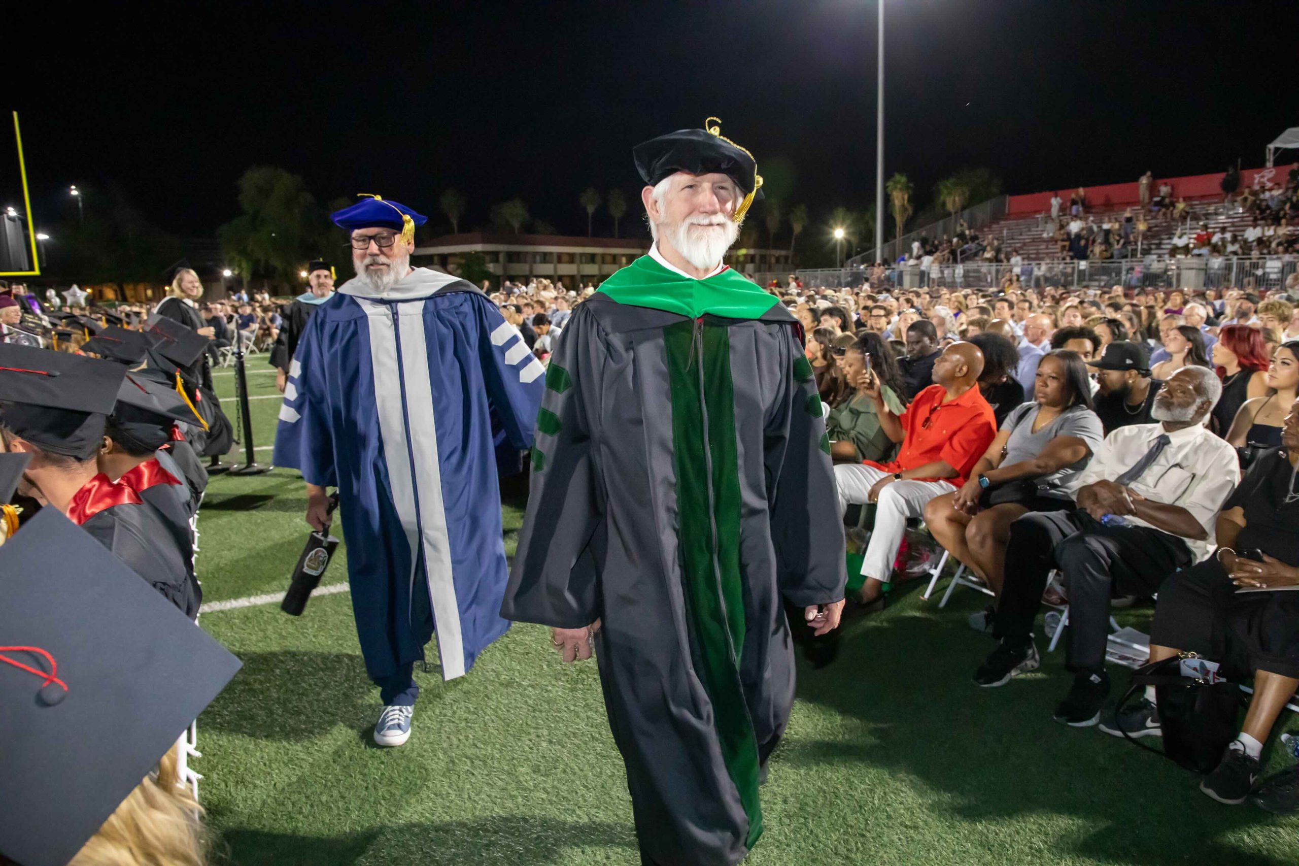 Graduates in academic regalia walk through a stadium field at night while seated spectators watch from the stands.