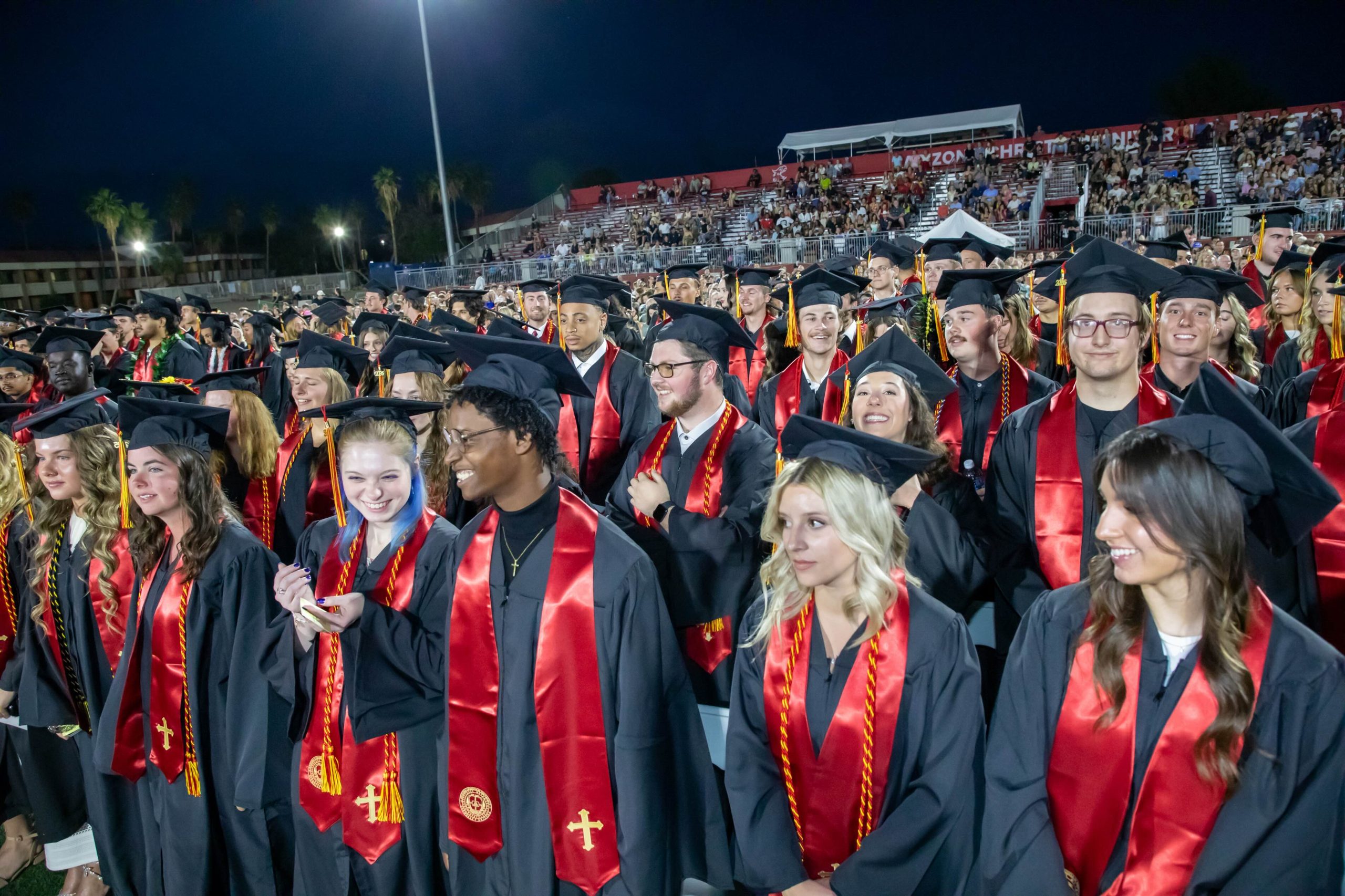 Crowd of graduates in black gowns and red stoles at a nighttime stadium ceremony with cheering audience in stands behind them.