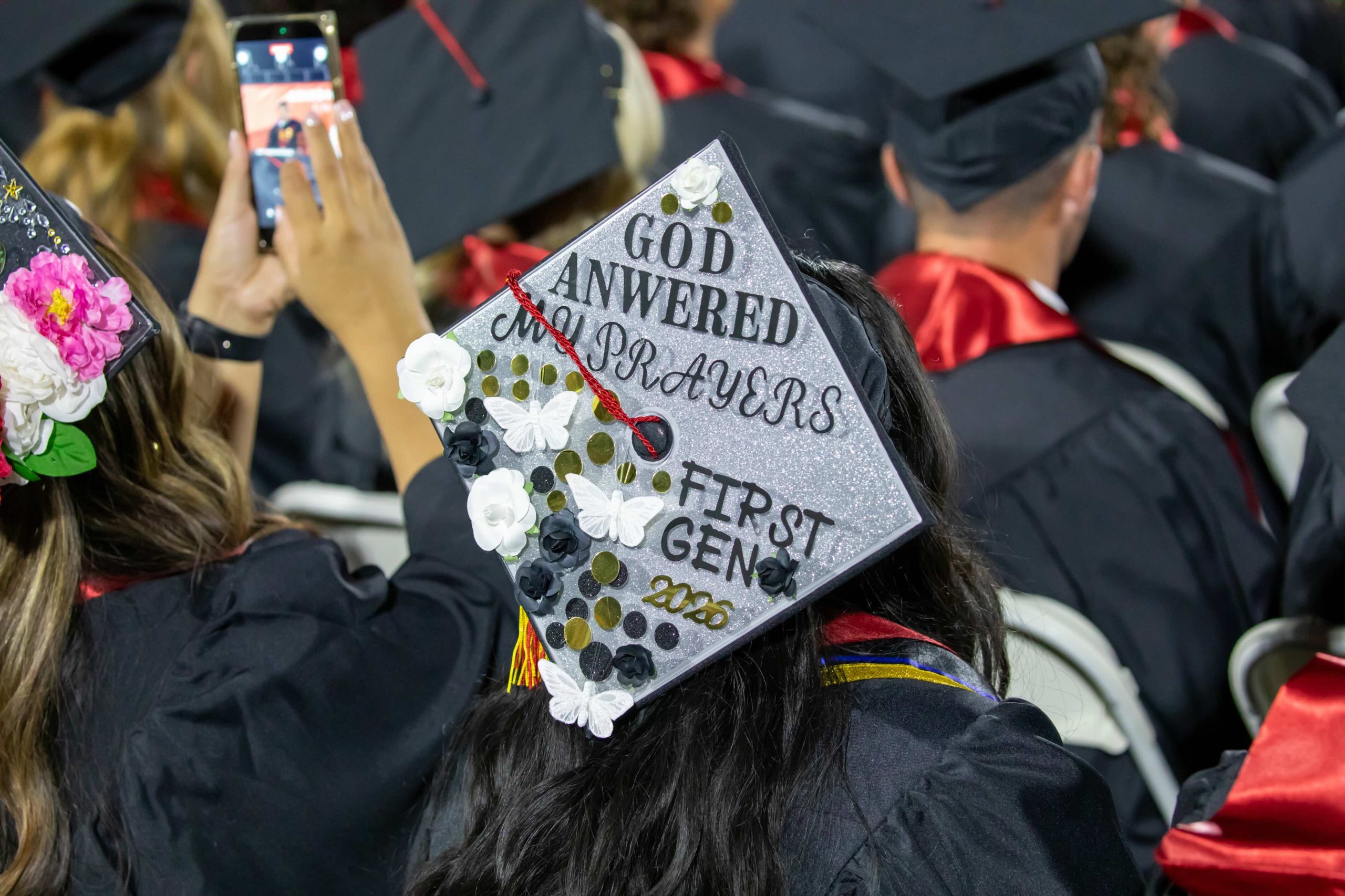 Graduation cap decorated with white flowers, butterflies, and glitter; reads "God answered my prayers first gen" at a ceremony backdrop.