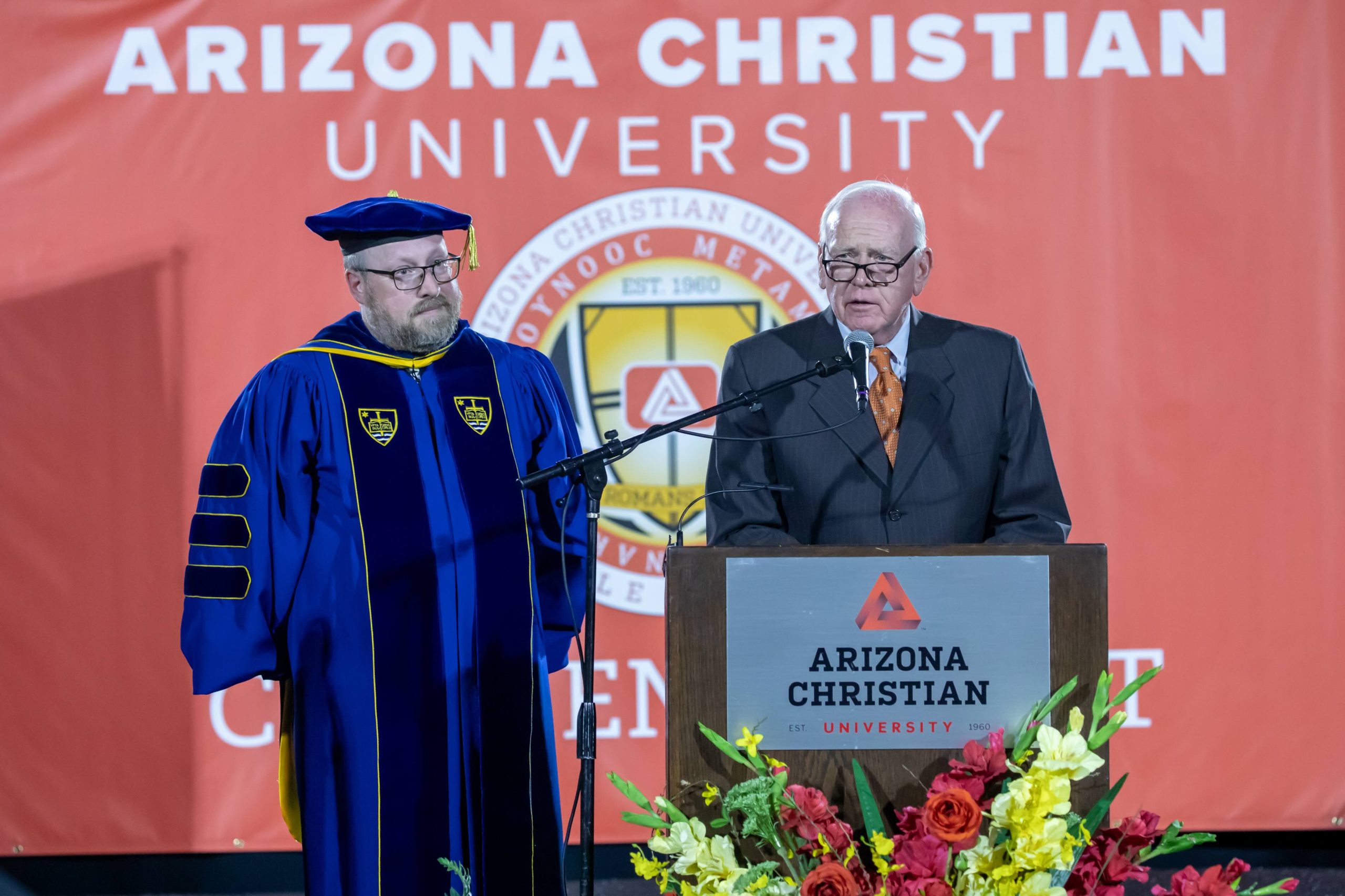 Two men on a stage at Arizona Christian University: a faculty member in royal blue doctoral regalia beside a suited speaker at a podium, with an orange university backdrop and floral arrangements in front.