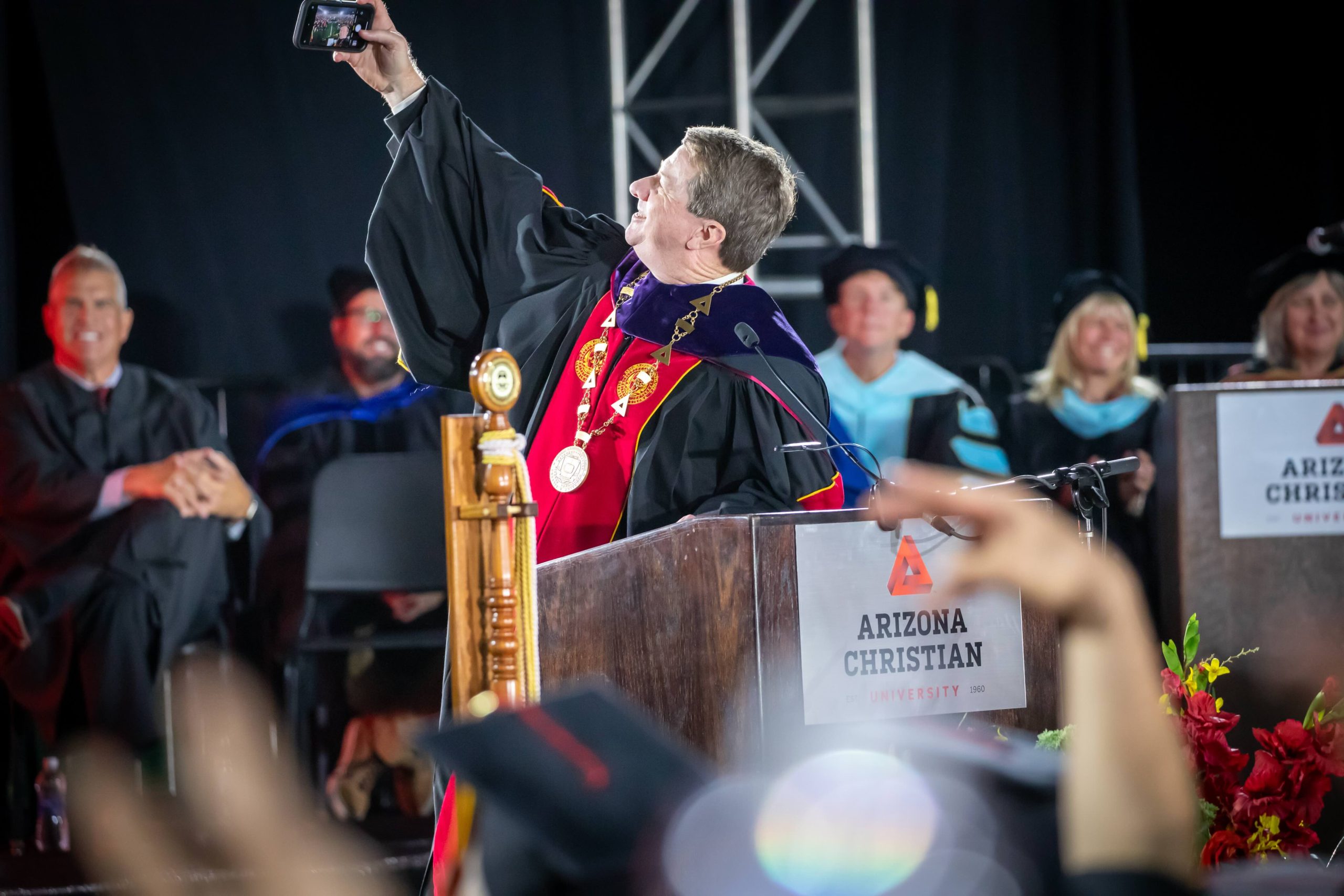 Graduate in gown and regalia takes a selfie at a podium during a ceremony on stage (Arizona Christian University)