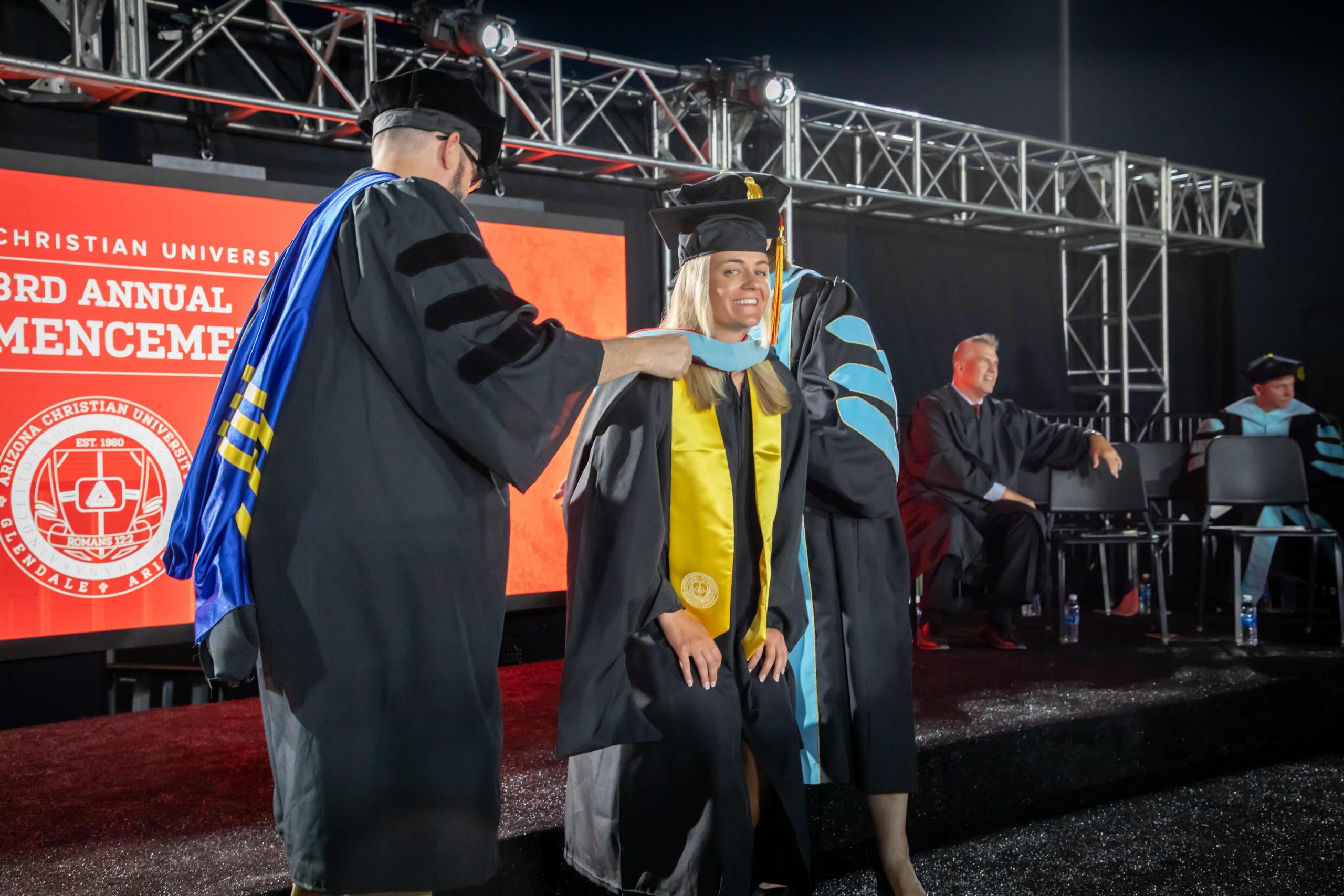 Graduate smiles as faculty places a blue-and-gold hood on her during a nighttime university commencement ceremony on stage with a red backdrop