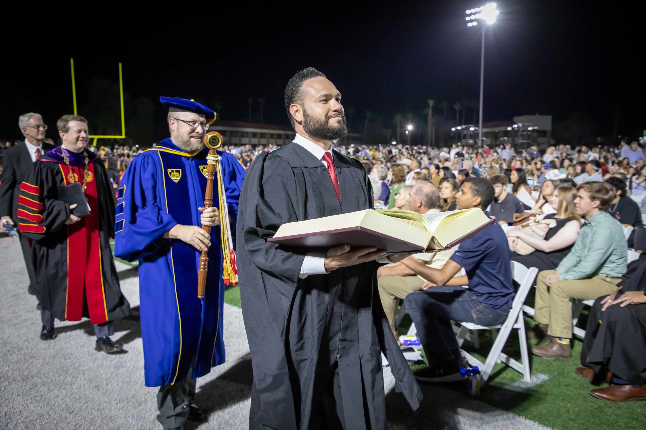 Graduate in black cap and gown holds an open book on stage during nighttime ceremony, with faculty in gowns nearby and a crowded audience in the background.
