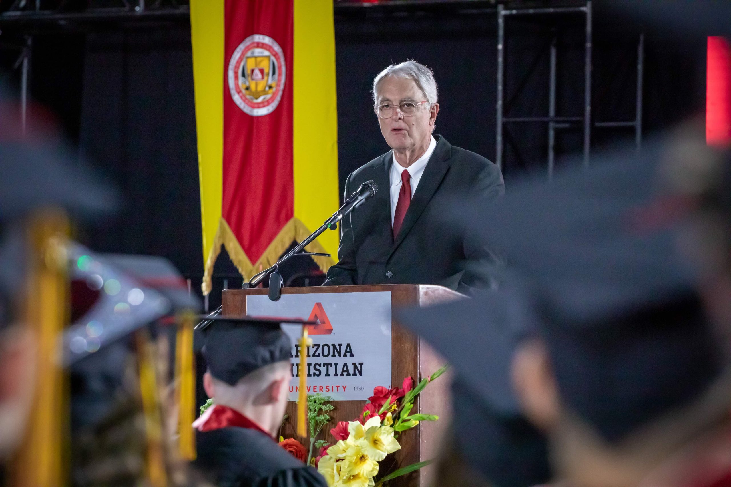 Older man in a dark suit delivering a speech at a podium during a graduation ceremony, with a red-and-yellow banner behind him and blurred graduates in the foreground.