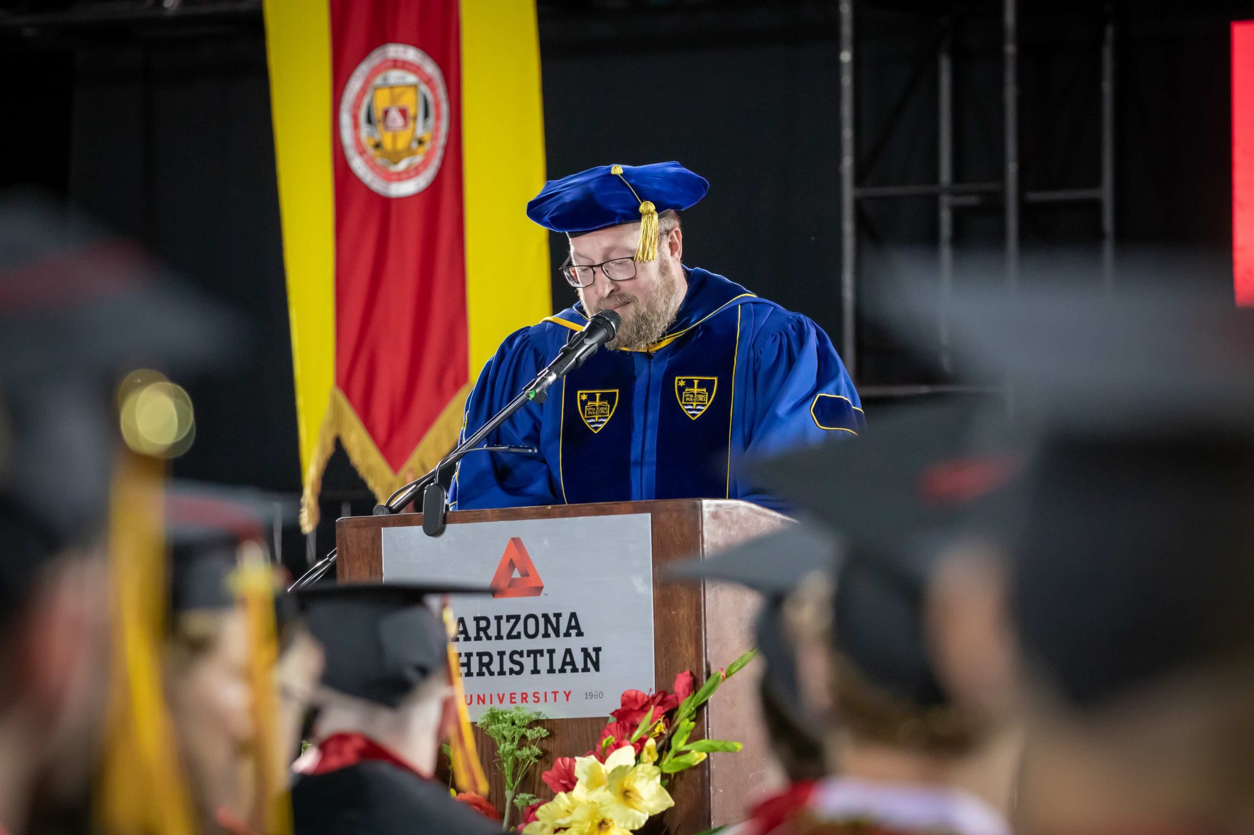 Man in a blue graduation gown and cap with a gold tassel speaks at a podium, with an Arizona Christian University banner behind him.