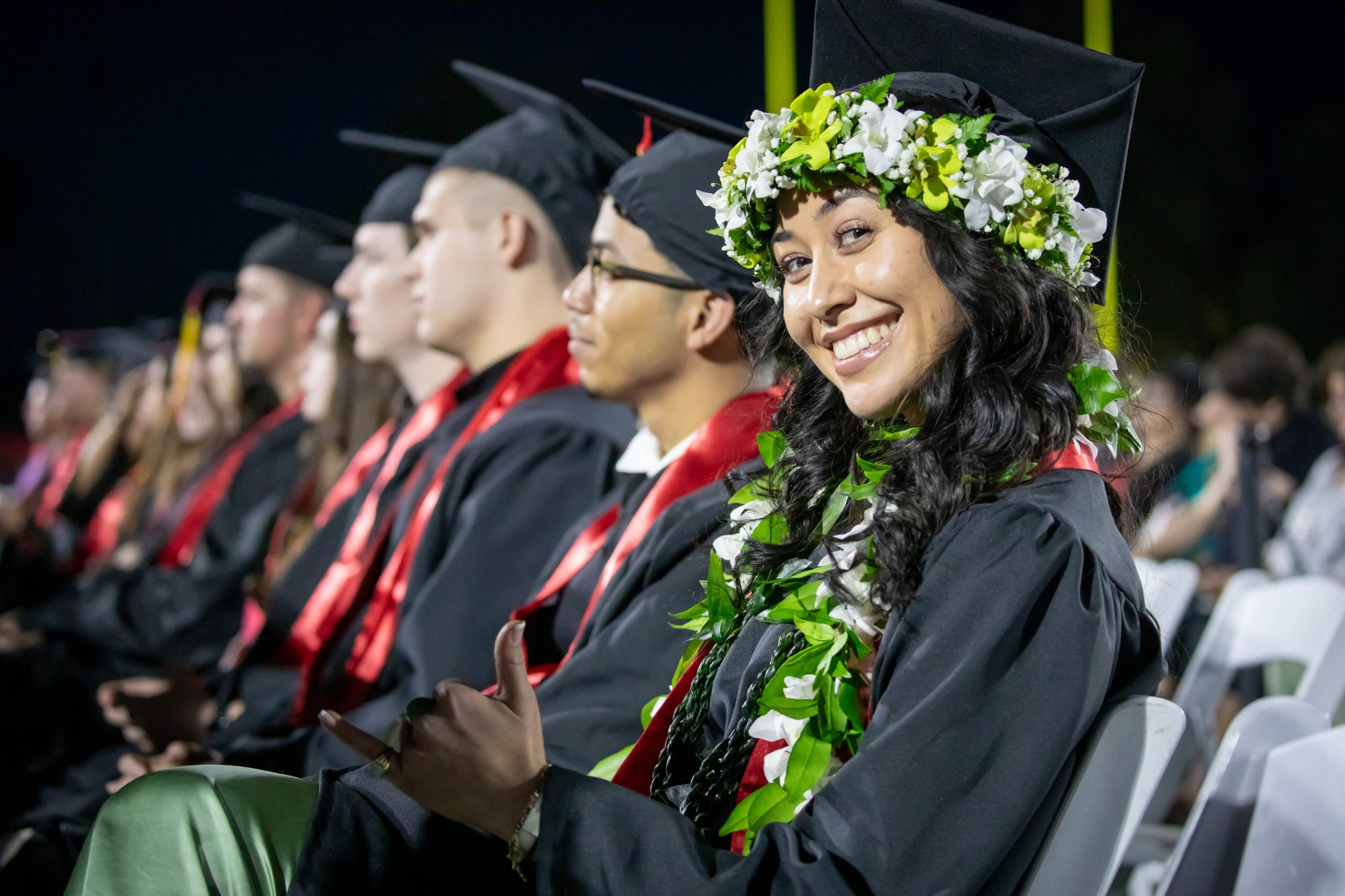 Smiling graduate in black cap and gown with a green and white leis, seated among fellow graduates at night graduation ceremony