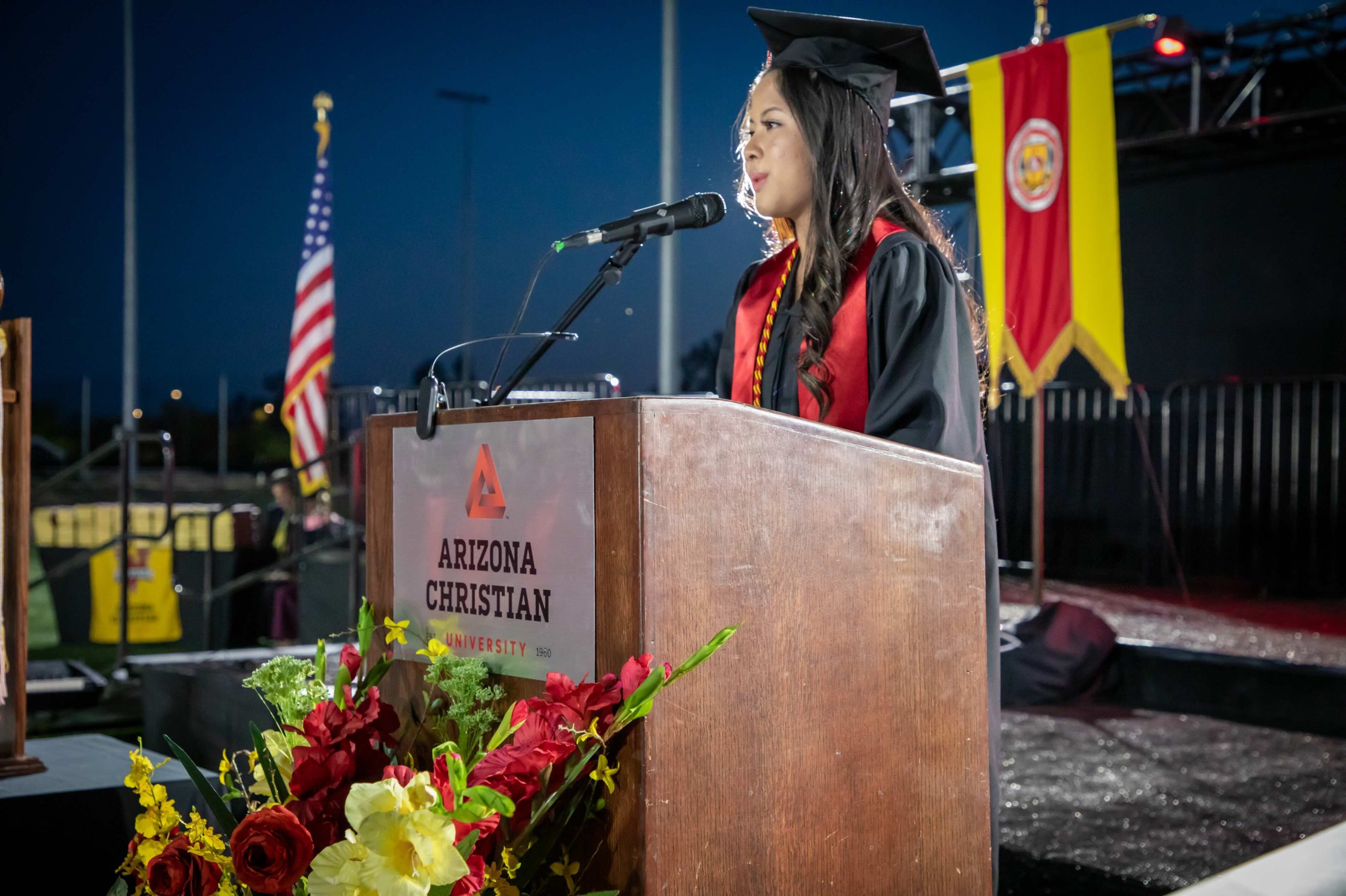 Female graduate in cap and gown speaks at podium during an outdoor graduation ceremony; Arizona Christian University logo on lectern with colorful flowers in foreground.
