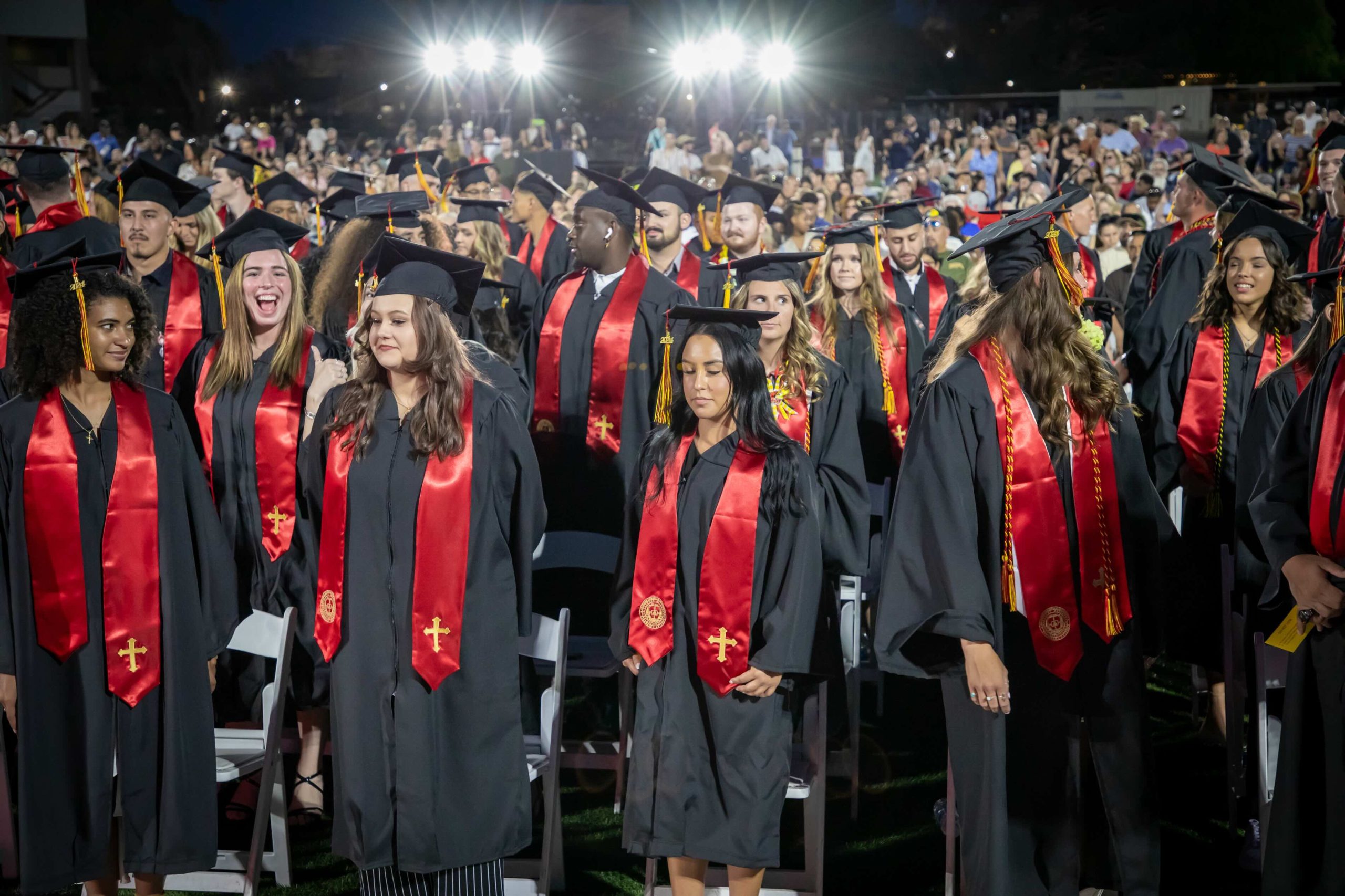 Outdoor graduation ceremony with graduates in caps and gowns, red stoles, under bright stadium lights at night.