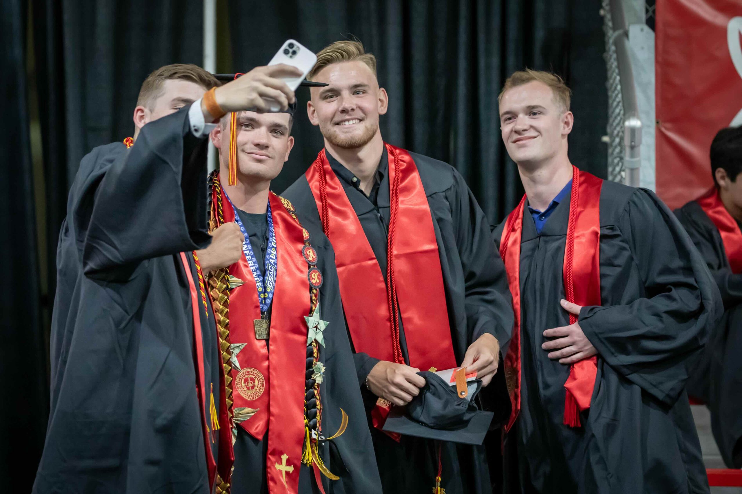 Three graduates in black gowns with red stoles pose for a selfie on stage, smiling as one holds up a smartphone.