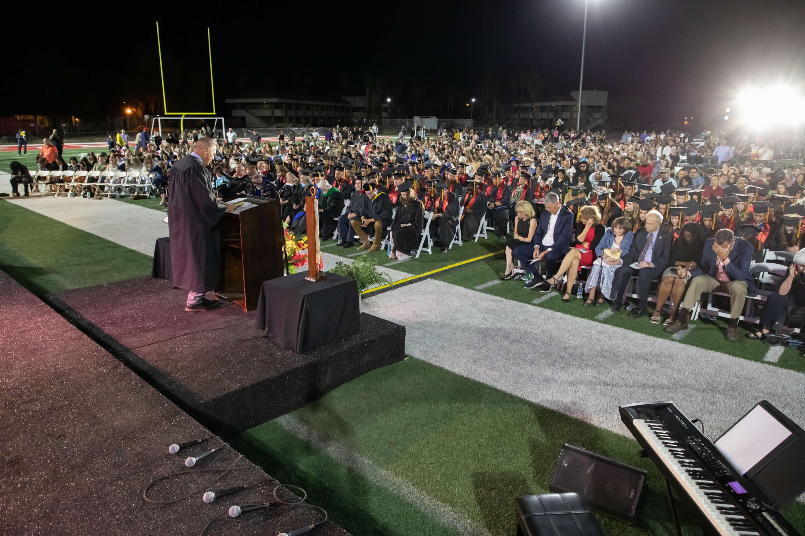Nighttime graduation ceremony on a football field: graduates in caps and gowns sit in rows as a speaker addresses them at the podium on a decorated stage.