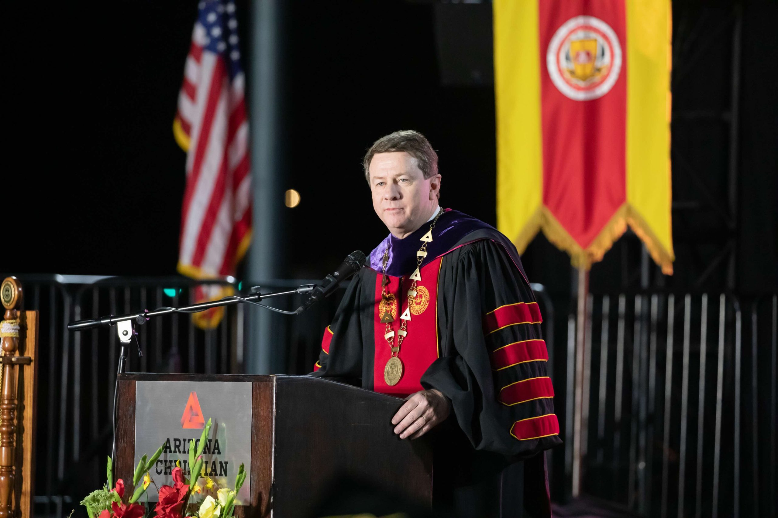 Man in ceremonial robes delivering a speech at a podium, with American flag and a university banner behind him, during a formal event with flowers nearby.