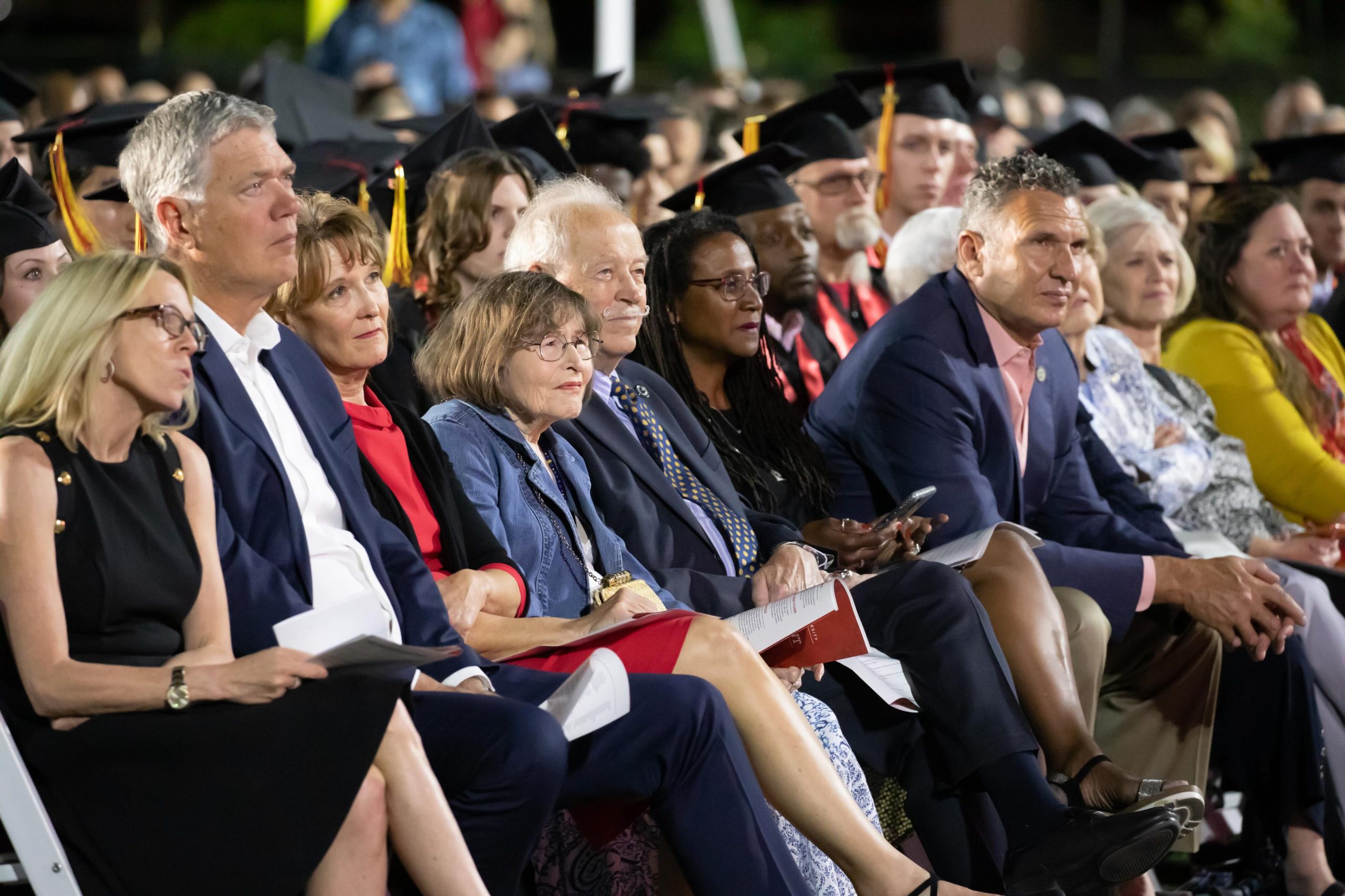 Audience seated at a graduation ceremony, many wearing caps and gowns and holding programs.