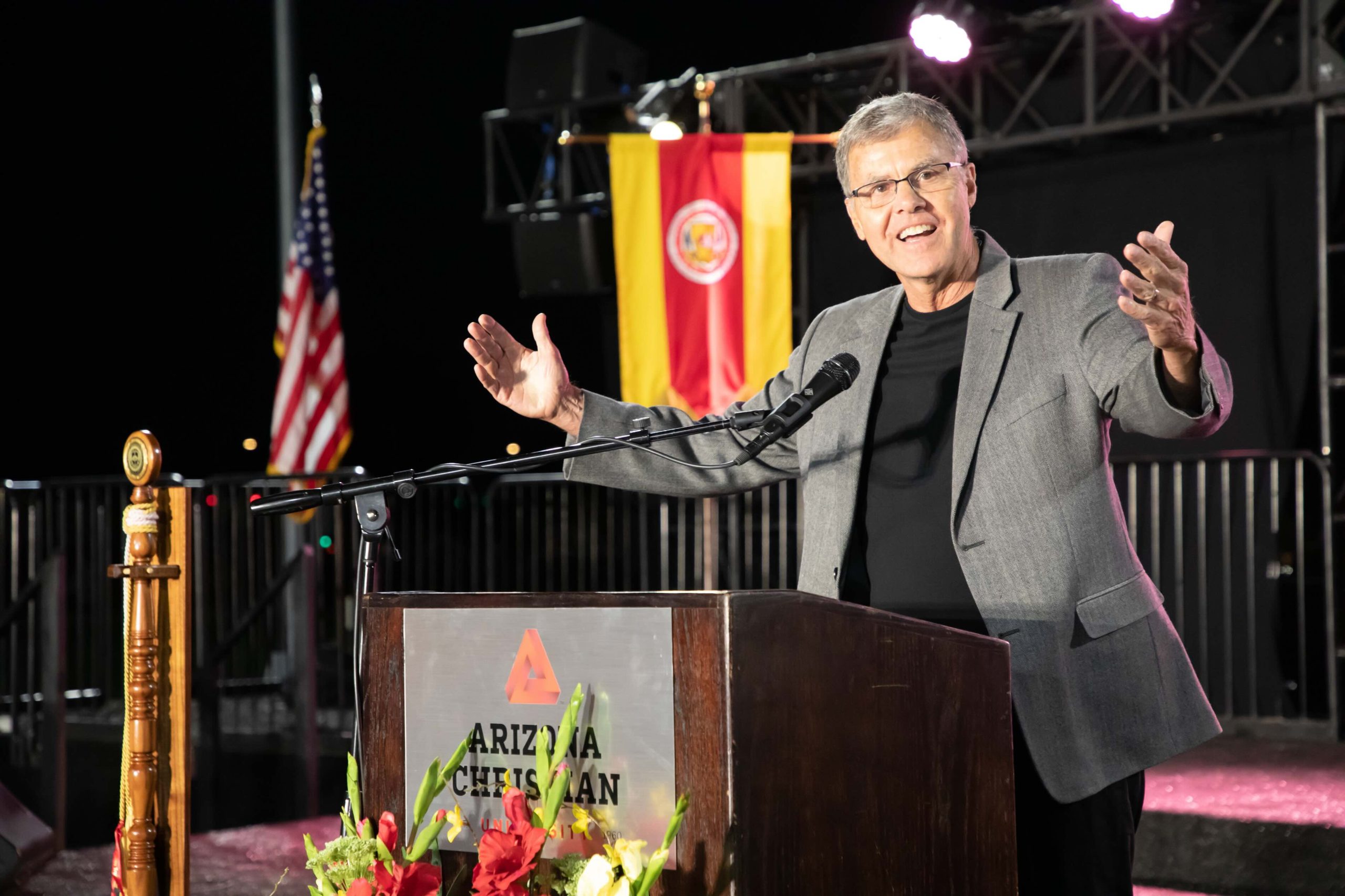 Speaker at a podium during a night outdoor event, arms outstretched, with American and red-yellow flags in the background.