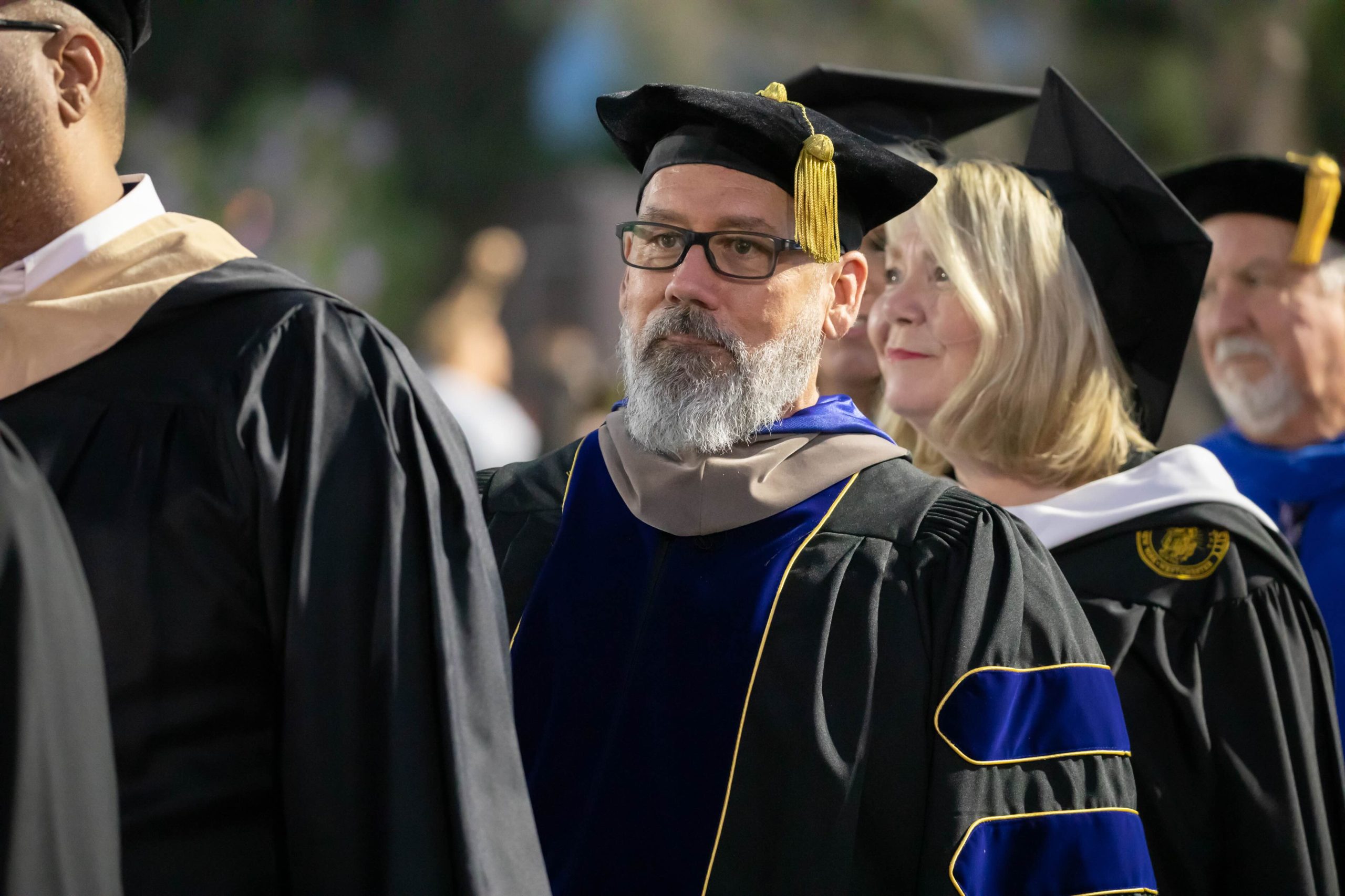 Smiling graduate in a black cap and gown with a gold tassel, among fellow graduates at a ceremony.