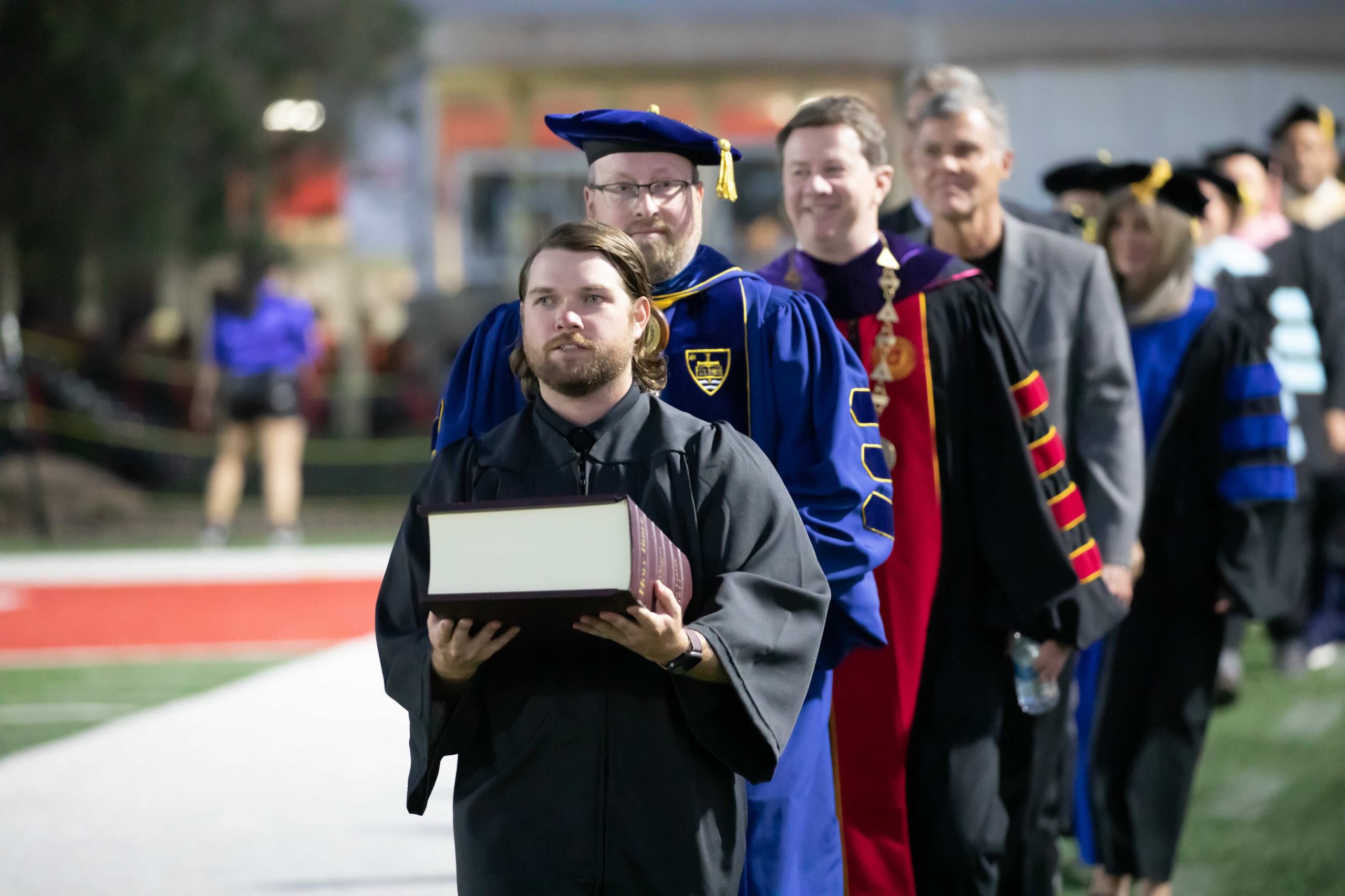 Graduate in a black gown carrying a diploma during a graduation procession with faculty in colorful regalia behind him