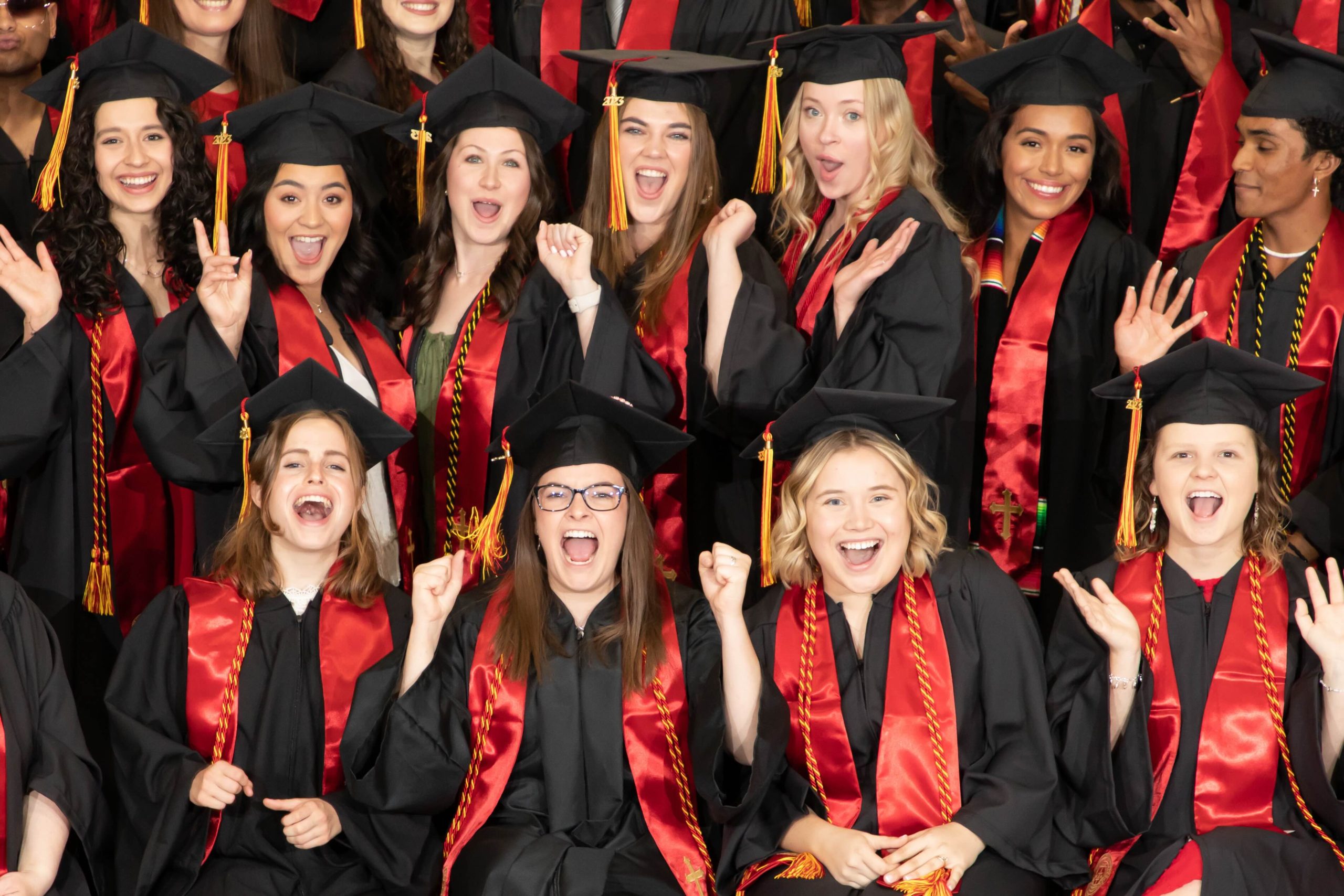 Group of diverse graduates in black gowns and red sashes cheering and posing for a photo.