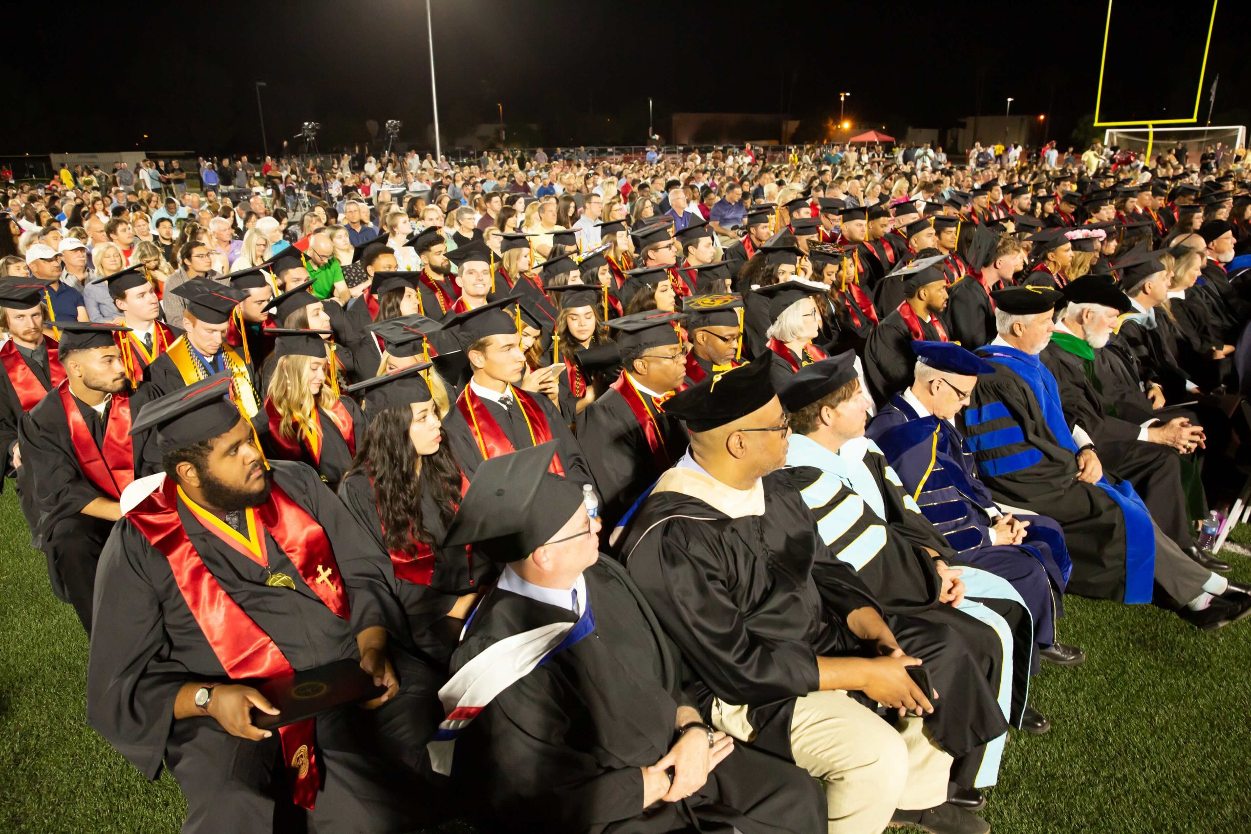 Large crowd of graduates in caps and gowns seated outdoors at night during a commencement ceremony.