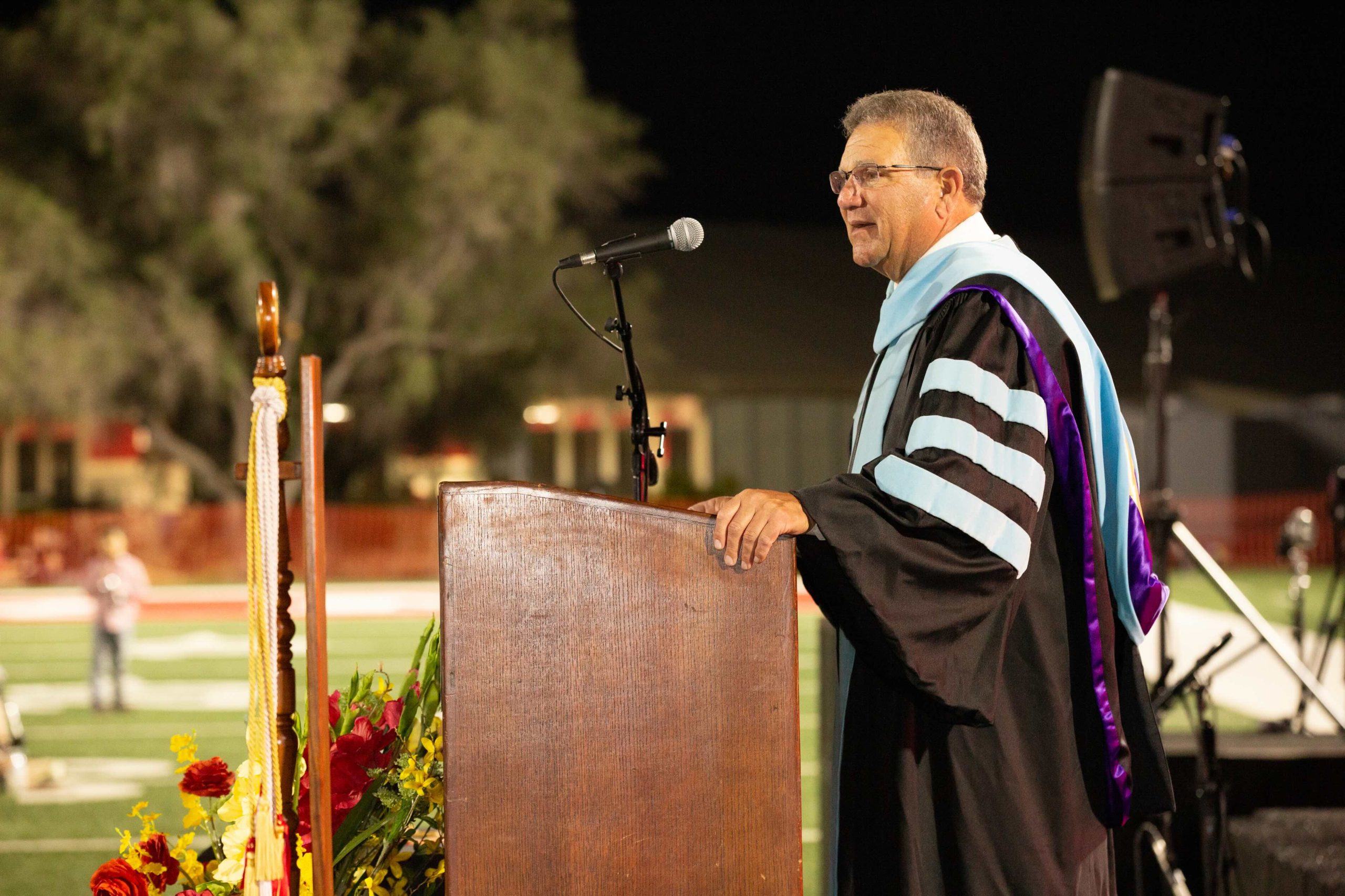 Graduation speaker in black robes with light blue and purple trim delivering a speech at a wooden podium with a microphone, flowers in the foreground.