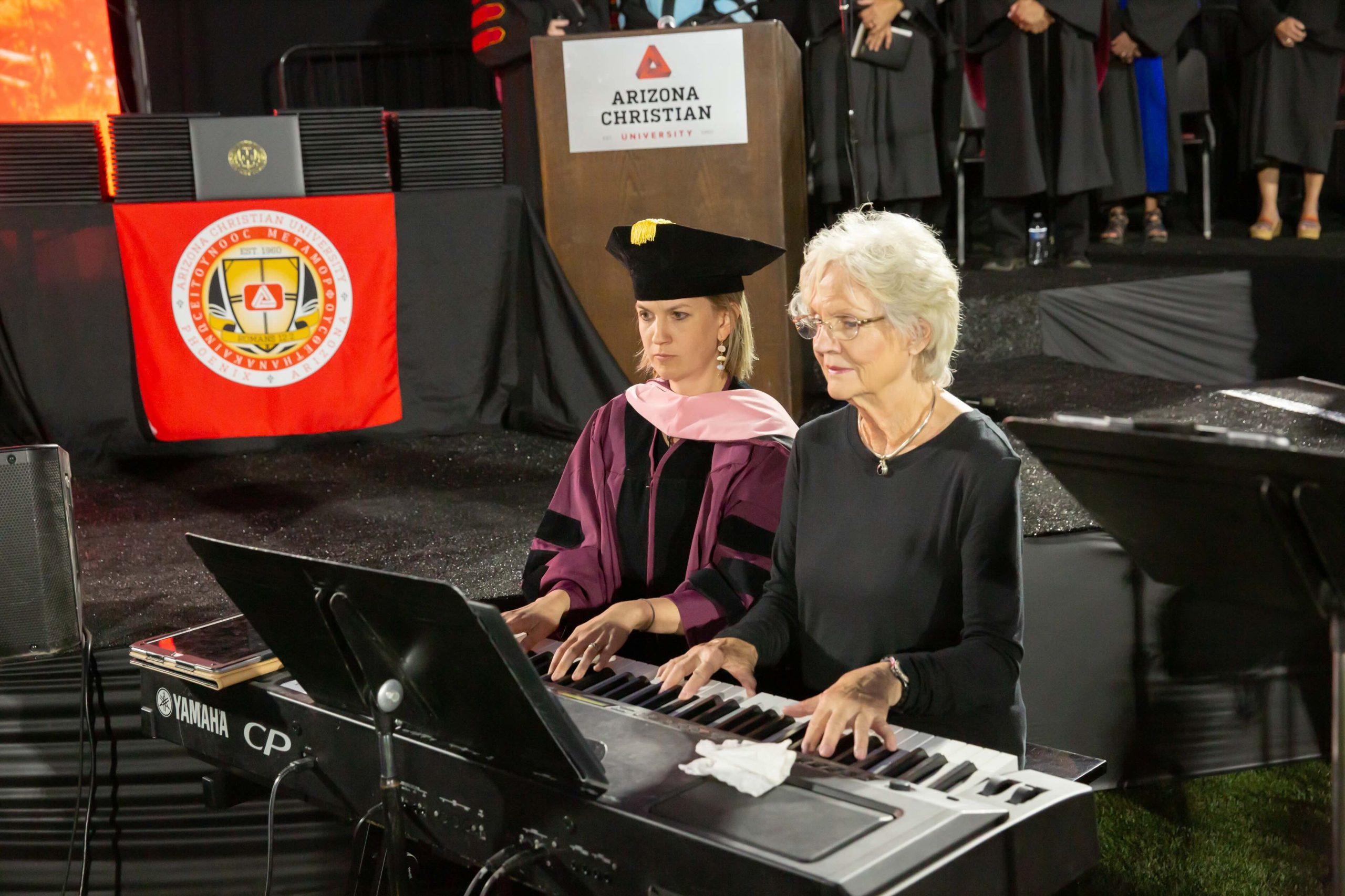 Two women in graduation regalia play a Yamaha CP keyboard on stage at an Arizona Christian University ceremony.