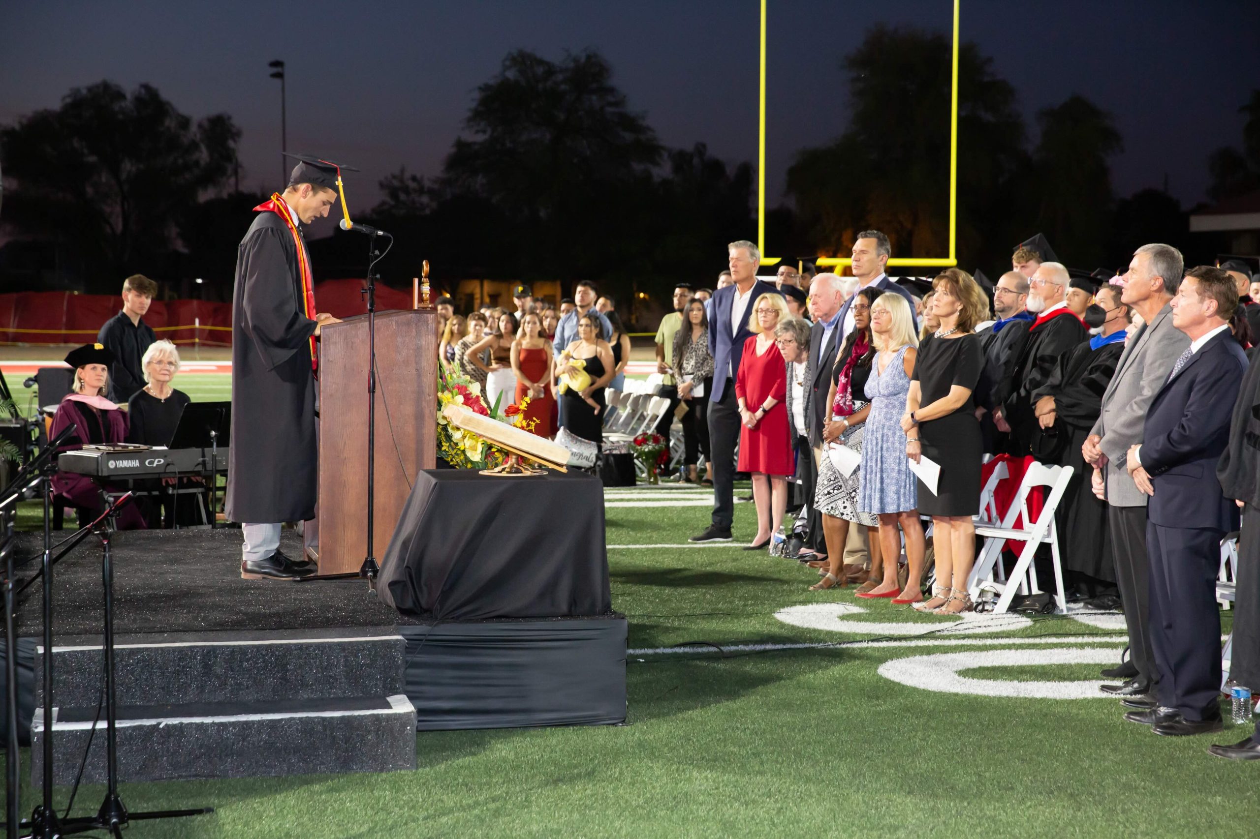 Graduate in cap and gown speaks at a podium during an outdoor graduation ceremony as the audience stands behind on the football field at dusk/nighttime.