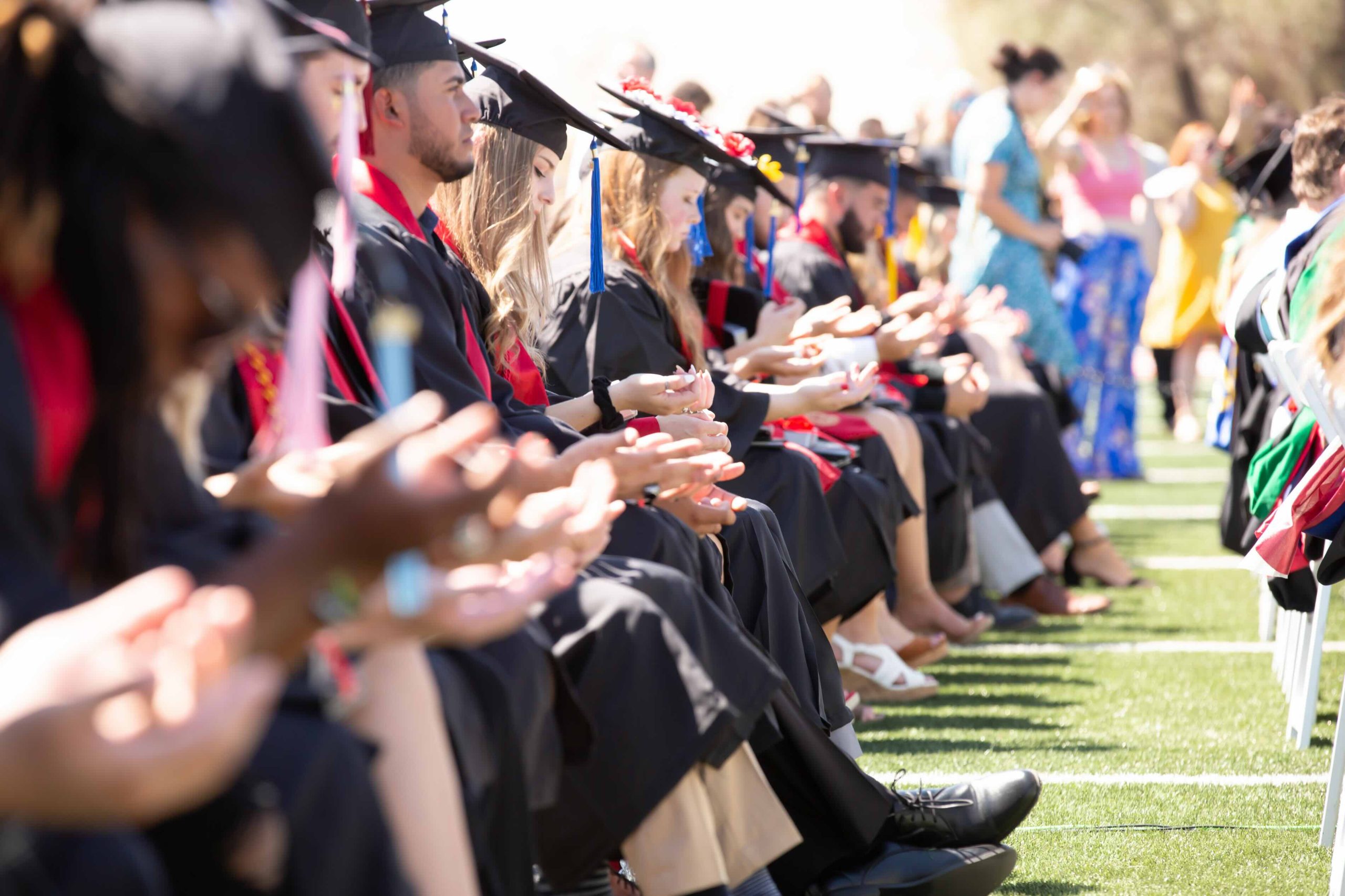 Graduates in caps and gowns sit in a row outdoors, clapping during a graduation ceremony.