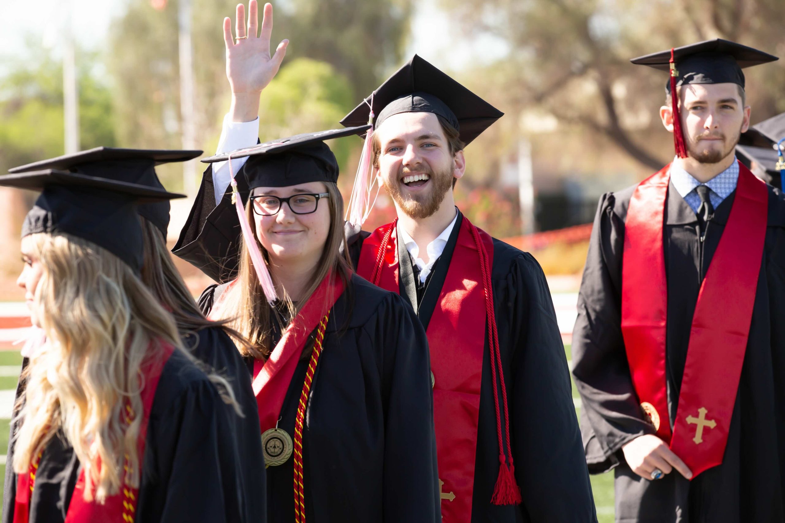 Group of graduates in black gowns and mortarboards at an outdoor ceremony, one waving and smiling at the camera.