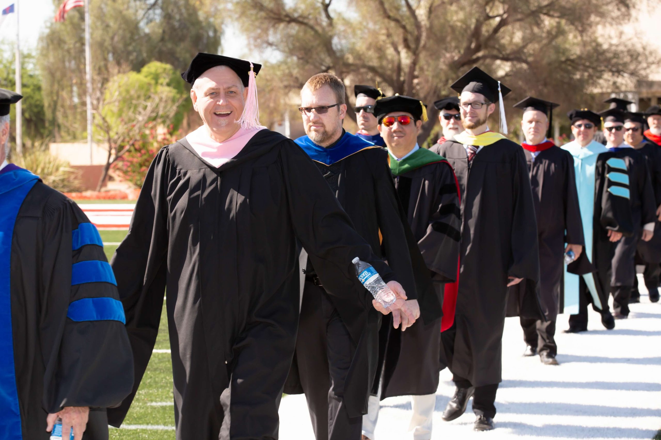 Line of graduates in caps and gowns walking outdoors during a ceremony, sunlight and trees in the background, one person holding a water bottle in the foreground.
