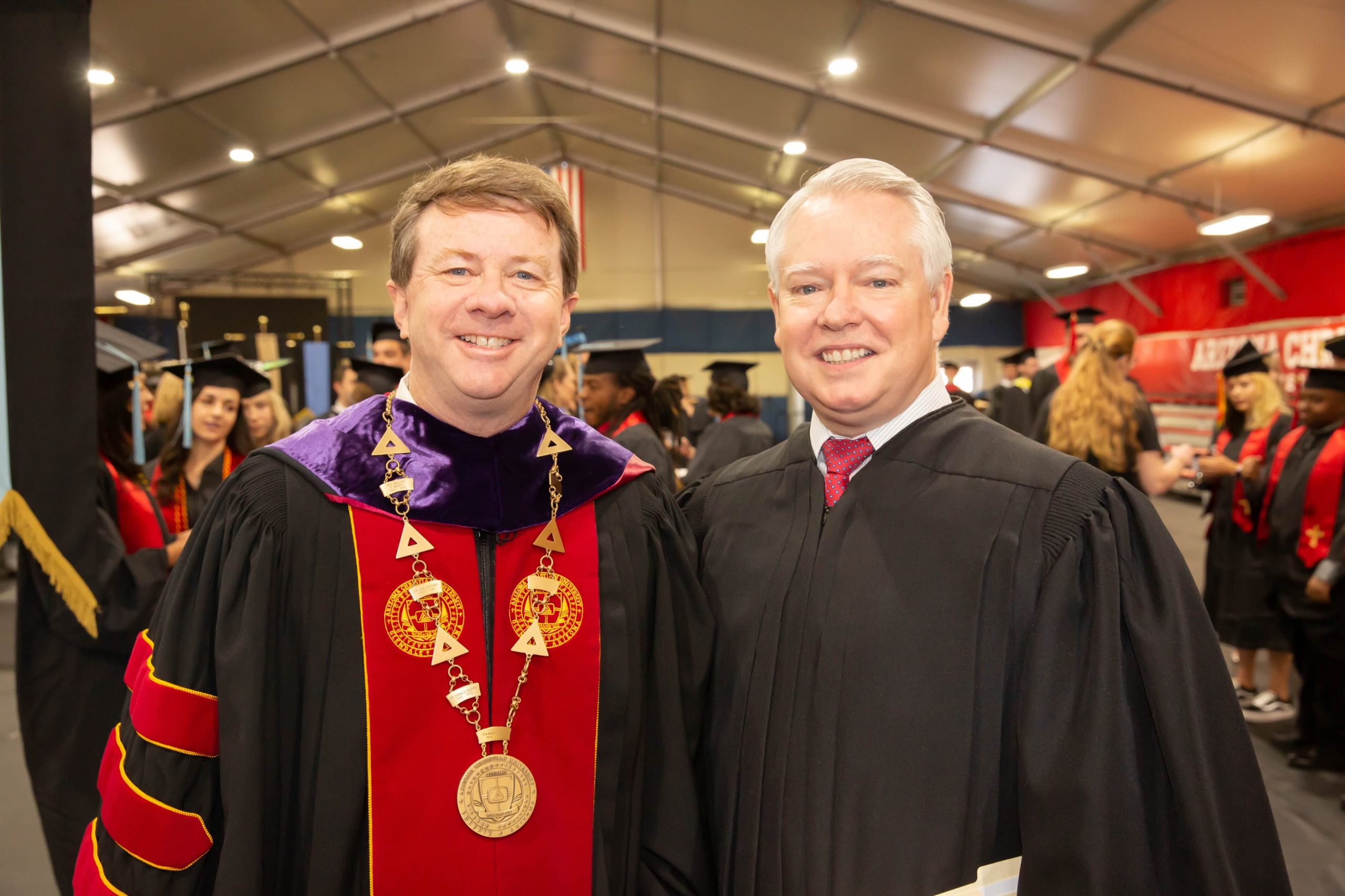 Two graduates in cap and gown posing for a photo at a graduation ceremony, one wearing a red and gold ceremonial stole and chain, the other in black regalia, smiling.