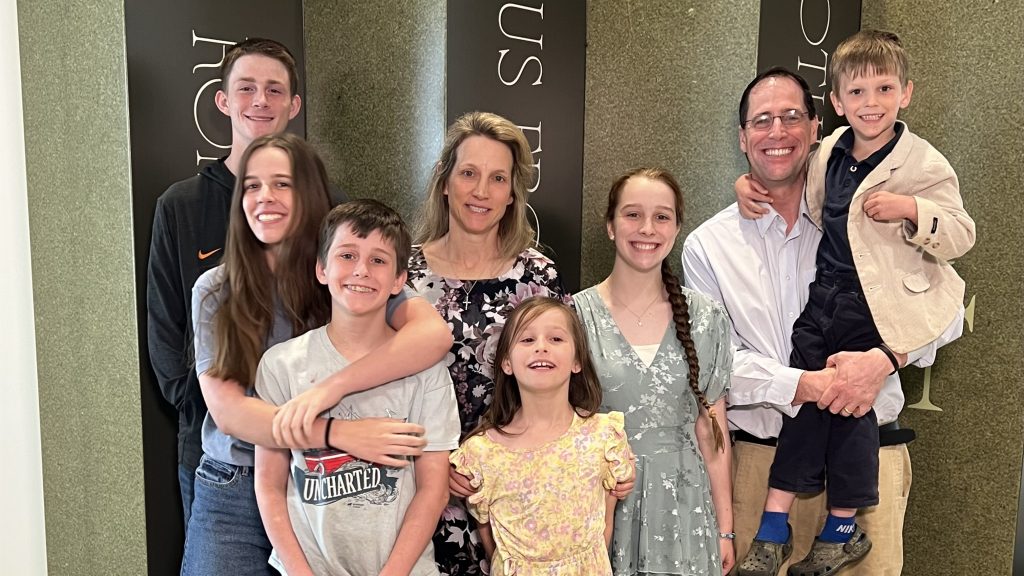 Large family group photo indoors; everyone smiles at the camera, with adults in back and children in front against a wall with vertical banners nearby.