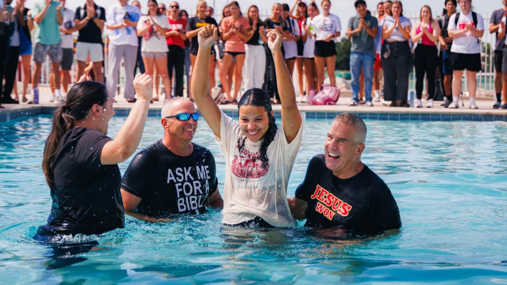 A girl raises her arms in celebration as she is baptized in a pool, with two men beside her and a cheering crowd on the pool deck behind them.