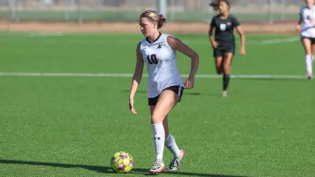 Female soccer player in a white jersey (number 16) dribbling the ball on a green field during a match, with teammates in the background.