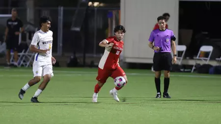 ACU Earns First Win Over Nelson - Arizona Christian University Red-team soccer player dribbles the ball past white-team opponents on a green turf while a purple-shirted referee watches nearby.