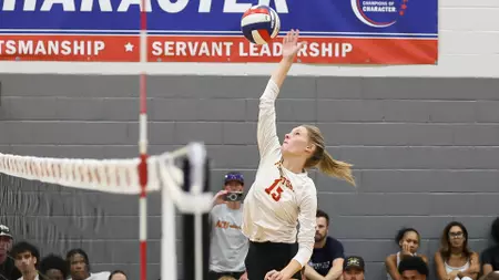 Female volleyball player in a white jersey (number 15) jumps to spike the ball over the net in a gym, with banners and spectators in the background.