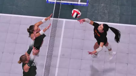 Overhead view of a volleyball rally: two blockers at the net on the left and a spiker on the right reaching for the ball.