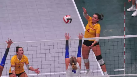 Women’s indoor volleyball match: two yellow-jersey players jump to block at the net while a teammate in white prepares to receive as the ball travels over.