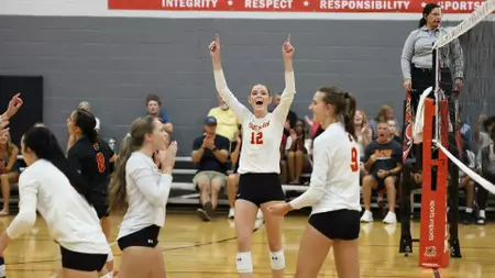 A volleyball player wearing number 12 raises both arms in celebration while teammates celebrate on the court behind her in a gymnasium.