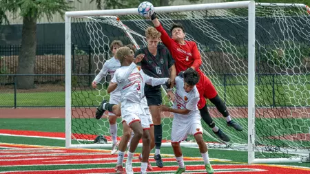 Soccer scene at a goal: players from white and red teams scramble for a header as the ball flies toward the net, with the goalkeeper in red mid-leap.