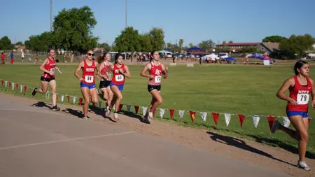 Female runners in red uniforms race along a sunlit track boundary on a grassy field, with spectators and tents in the background.
