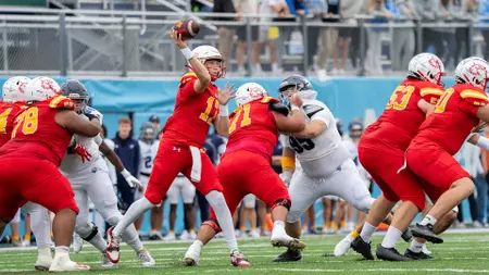 Quarterback in red jersey throws a pass while teammates block defenders on a football field during a game.