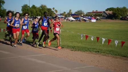 Group of runners in blue and red uniforms sprinting at the start of a track race on a sunny day, with pennant flags and spectators in the background.