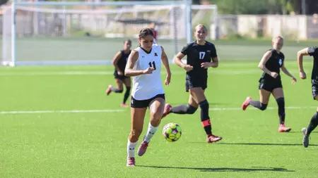 Female soccer player in white jersey #11 dribbles the ball down the field as defenders in black chase nearby on a sunny turf pitch.