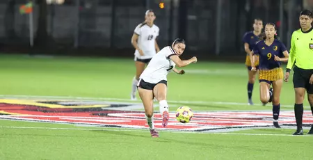 Female soccer player in a white jersey and black shorts kicks a ball on a turf field during a night match, with opponents and a referee nearby.