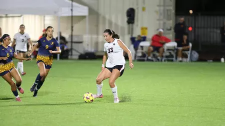 Female soccer player in white jersey #33 dribbles the ball on a green indoor field as defenders in blue-yellow chase behind her.
