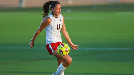 Female soccer player in a white jersey (number 11) and red shorts dribbling a bright yellow ball on a green field.