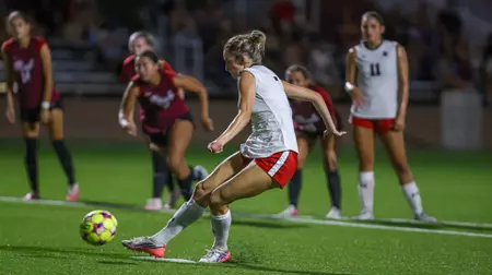 Female soccer player in white and red shorts kicking a ball during a nighttime match as opponents chase.